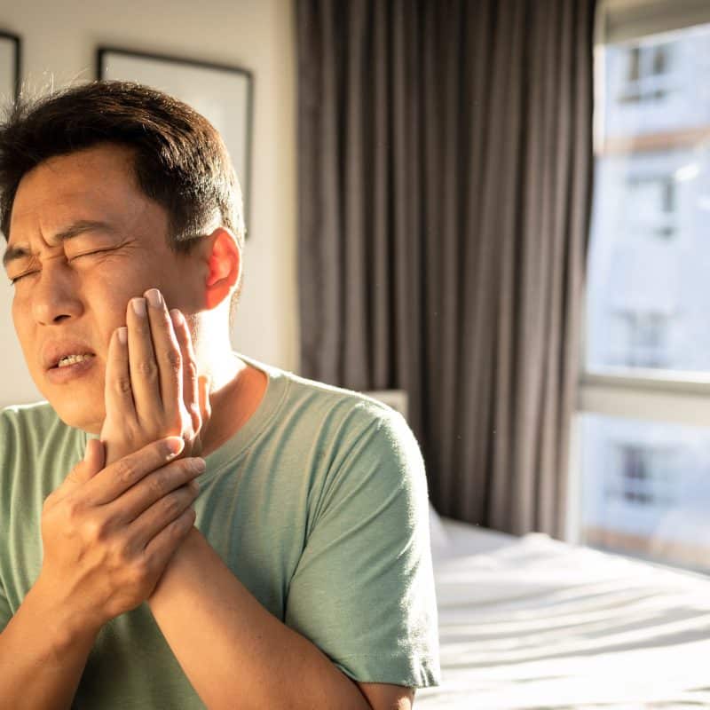 man sitting up in bed holding his jaw, extreme pain, possible TMJ sufferer