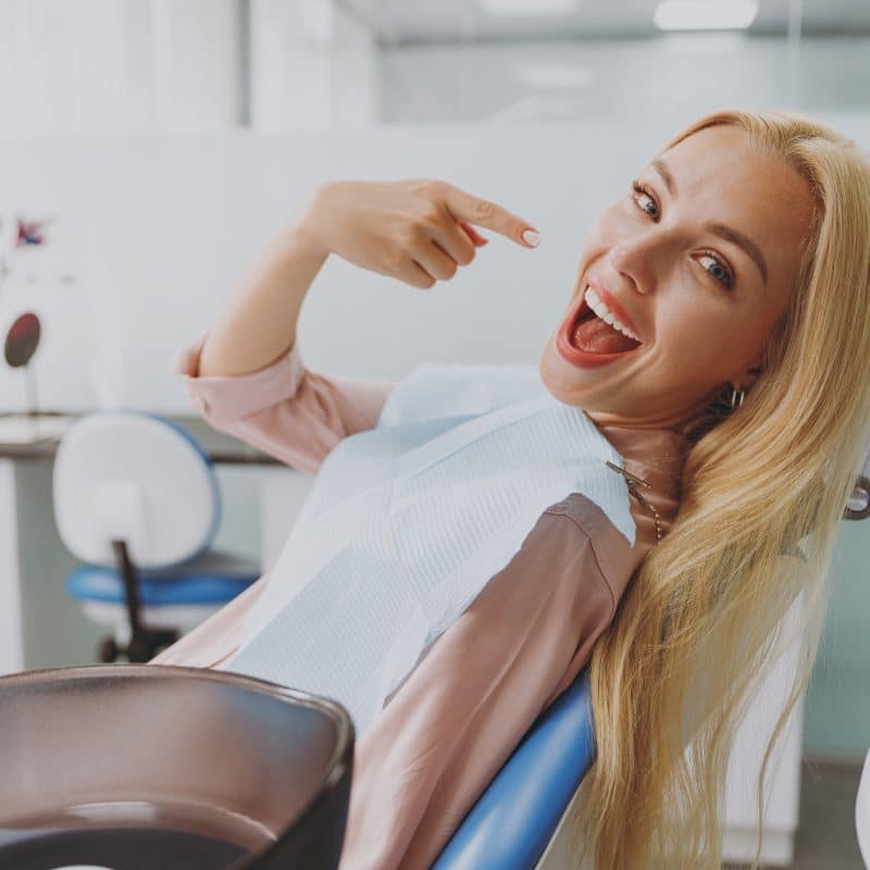 blonde haired woman sitting in a dental chair, pointing to her new smile